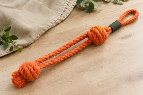 Jouet de traction Trixie en coton orange avec double corde et dragonne, posé sur un sol en bois clair.